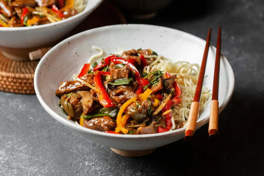 A vibrant stir-fry dish featuring tender beef strips, colorful bell peppers, and fresh herbs, served over a bed of noodles with chopsticks resting on the side.