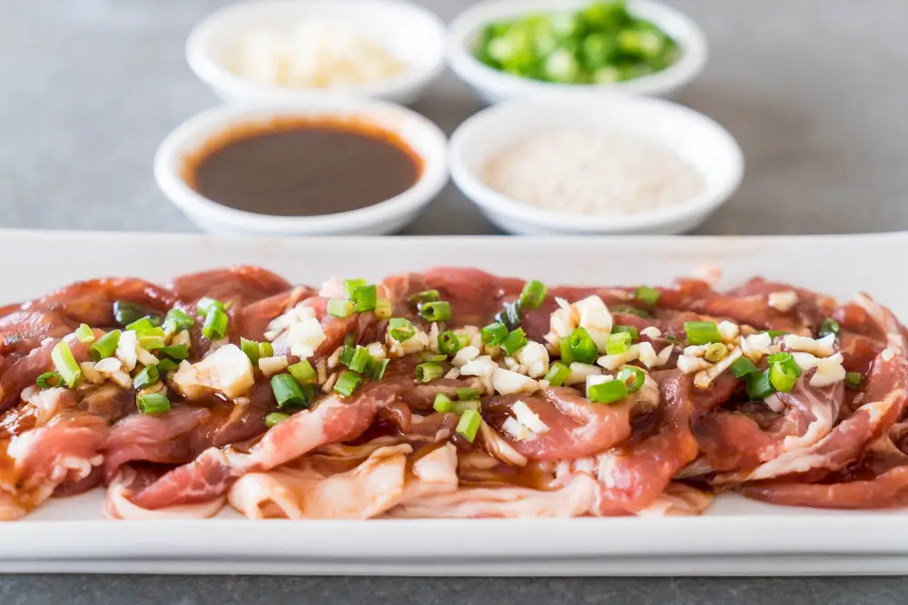 Thinly sliced beef arranged on a white plate, garnished with chopped green onions and garlic, with small bowls of sauces and condiments in the background.