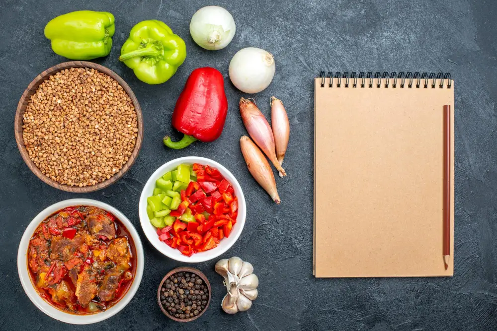 A variety of fresh ingredients for cooking, including green and red peppers, onions, shallots, garlic, buckwheat, and a bowl of mixed vegetables, arranged on a table with a notebook.