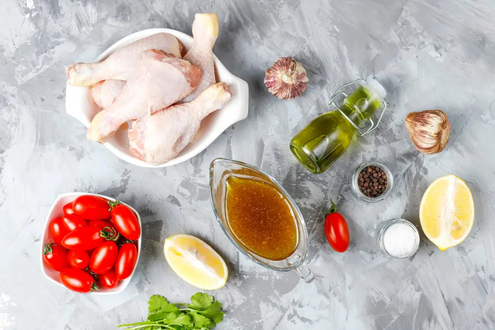 A top-down view of raw chicken drumsticks, cherry tomatoes, garlic, olive oil, lemon, spices, and marinade on a textured gray surface