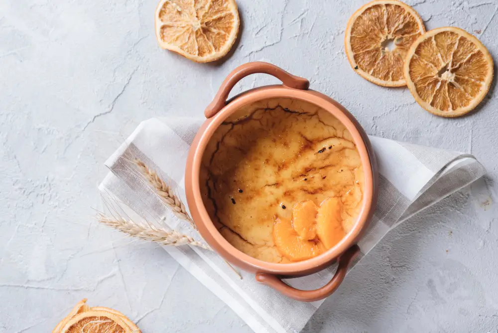 A close-up of a traditional Moroccan dessert, featuring a creamy, caramelized top layer, served in a rustic clay pot with dried orange slices as garnish.