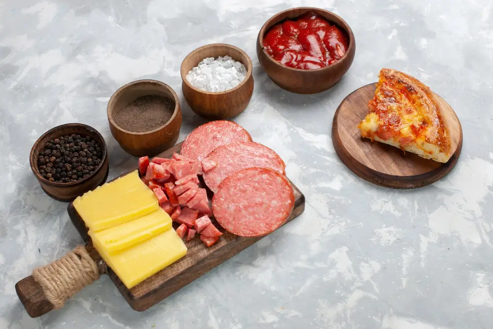 A wooden board with slices of cheese, salami, and diced ham, accompanied by bowls of pepper, salt, spices, and tomato sauce, with a slice of pizza on a wooden plate.