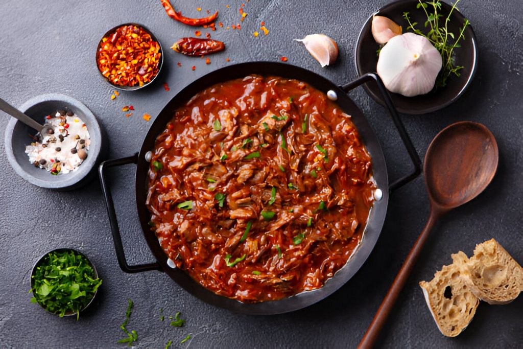 A pan of rich Beef Stew with tender meat, spices, and herbs, accompanied by garlic, chili flakes, and fresh bread.