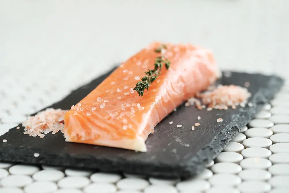 A fresh salmon fillet seasoned with salt and garnished with a sprig of thyme, placed on a dark slate board.