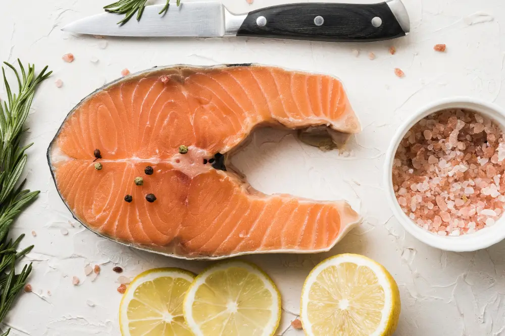 A raw salmon steak seasoned with pepper, accompanied by a knife, a bowl of pink salt, lemon slices, and a sprig of rosemary.