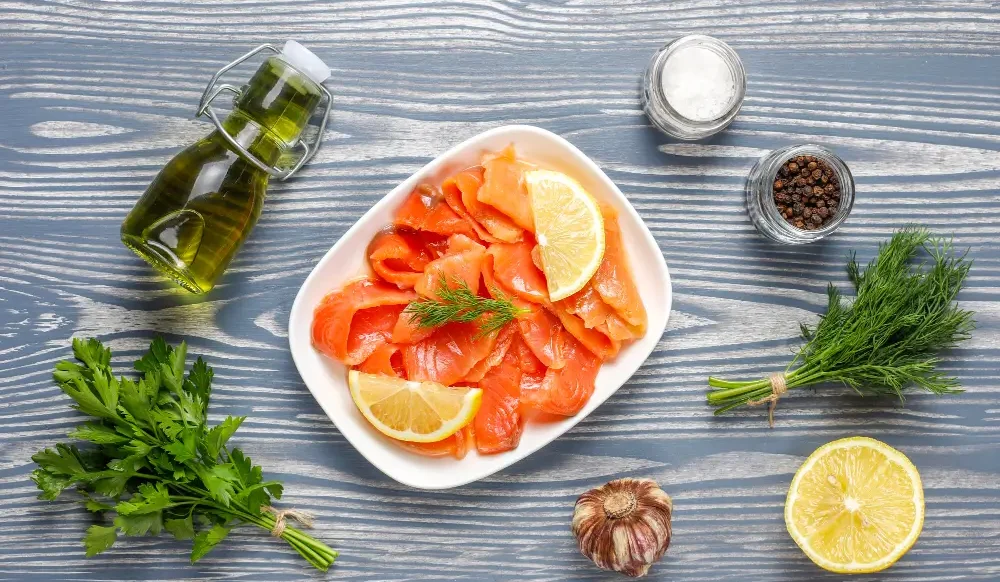 A plate of smoked salmon garnished with lemon slices and dill, surrounded by ingredients for a Smoked Salmon Brine Recipe including olive oil, salt, pepper, parsley, garlic, and lemon.