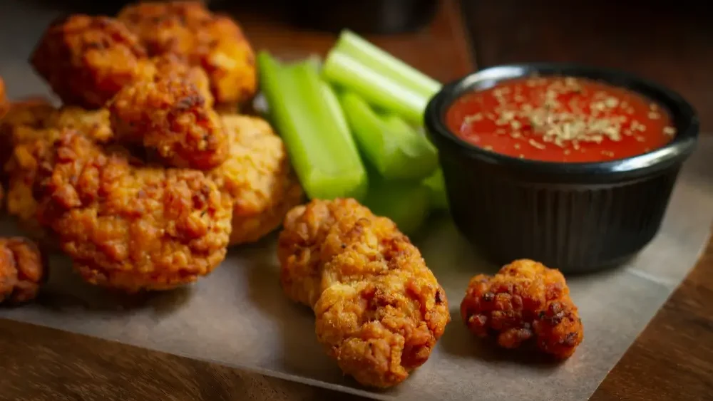 Crispy, golden-brown fried chicken bites served with fresh celery sticks and a bowl of red dipping sauce, garnished with herbs.